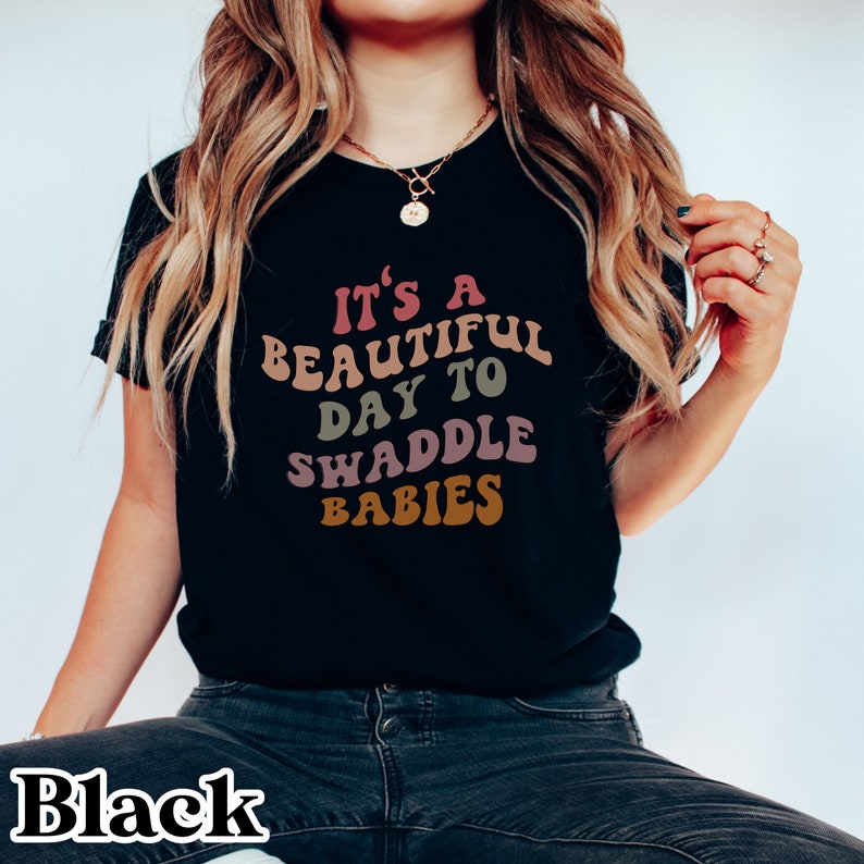 K&ouml;nnte beinhalten: Schwarzes T-Shirt mit dem Text "It's a beautiful day to swaddle babies" in bunter Retro-Schrift.