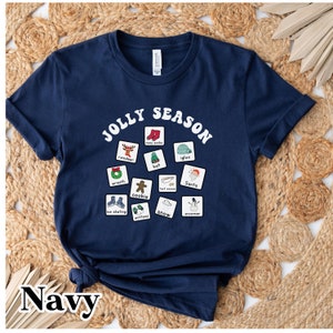 K&ouml;nnte beinhalten: Ein marineblaues T-Shirt mit einer wei&szlig;en Grafik der Worte "Jolly Season" in einer Retro-Schrift. Die Grafik enth&auml;lt auch verschiedene winterliche Symbole, wie zum Beispiel ein Rentier, einen Schneemann, einen Hut und einen Kranz.