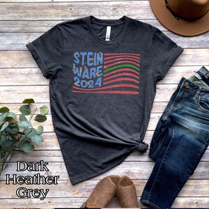 May include: Dark heather grey t-shirt with a distressed American flag design and the text "STEIN WARE 2024" in blue lettering.