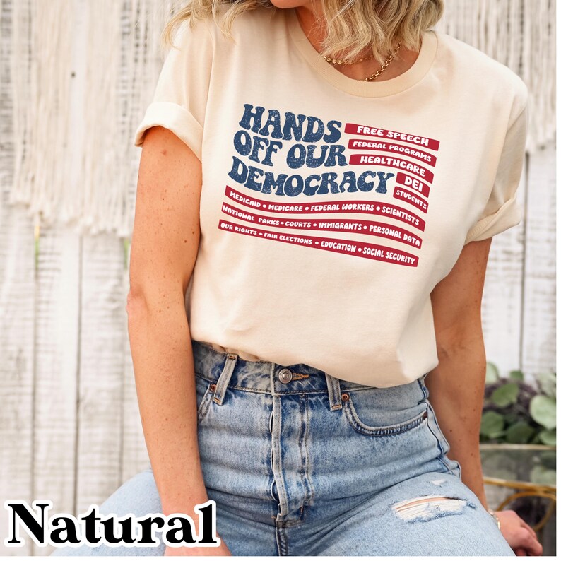 May include: A beige t-shirt with a red, white, and blue American flag design. The text "Hands Off Our Democracy" is printed in blue on the flag. The text "Free Speech, Federal Programs, Healthcare, DEI, Students, Medicaid, Medicare, Federal Workers, Scientists, National Parks, Courts, Immigrants, Personal Data, Our Rights, Fair Elections, Education, Social Security" is printed in black below the flag.