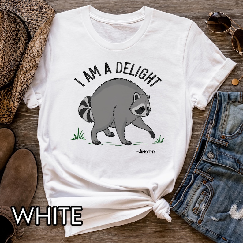 May include: A white short-sleeve t-shirt featuring a graphic of a grey raccoon standing on green grass. Above the raccoon, the text reads I AM A DELIGHT in black capital letters. Below the raccoon, the name -JIMOTHY is printed in black.