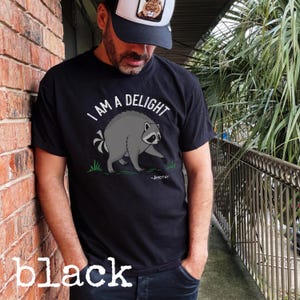 May include: A black short-sleeved t-shirt featuring a graphic of a grey raccoon standing on green grass. Above the raccoon, the text reads "I AM A DELIGHT" in white capital letters, and below the raccoon, the text reads "-JIMOTHY" in white.