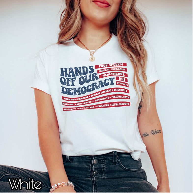 May include: White t-shirt with a blue and red graphic that says "Hands Off Our Democracy" with a list of items under it, including "Free Speech", "Federal Programs", "Healthcare", "DEI", "Students", "Medicaid", "Medicare", "Federal Workers", "Scientists", "National Parks", "Courts", "Immigrants", "Personal Data", "Our Rights", "Fair Elections", "Education", and "Social Security".