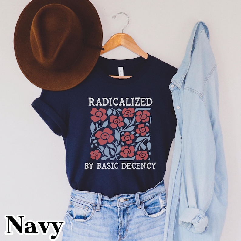 May include: Navy blue t-shirt with the text "RADICALIZED BY BASIC DECENCY" above a floral design in red and blue. A brown hat hangs above the shirt, and a light blue denim shirt is draped on the right.