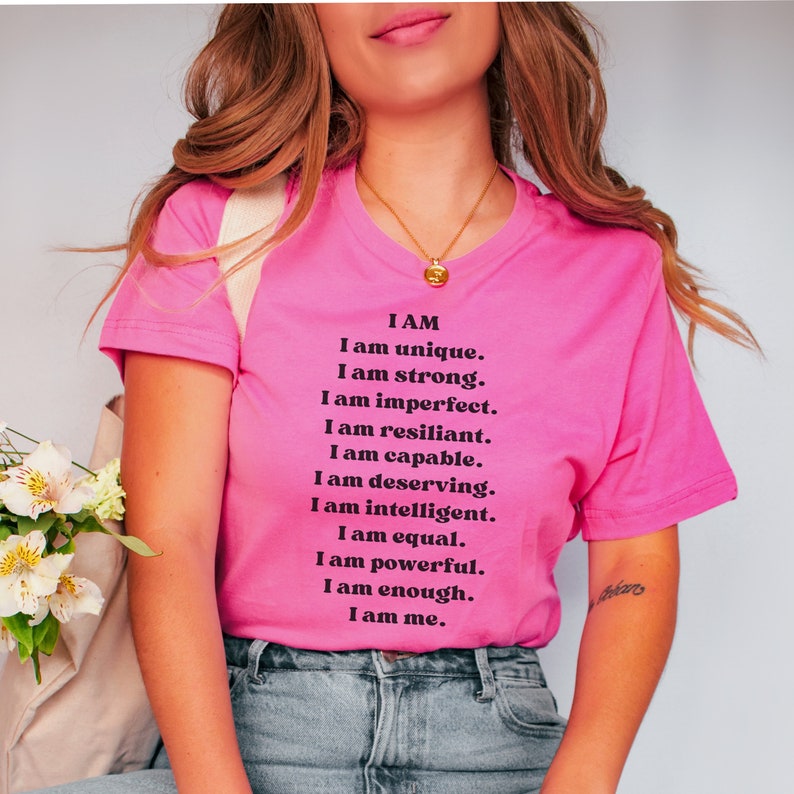 May include: A pink t-shirt with a black text message that reads "I AM I am unique. I am strong. I am imperfect. I am resilient. I am capable. I am deserving. I am intelligent. I am equal. I am powerful. I am enough. I am me."