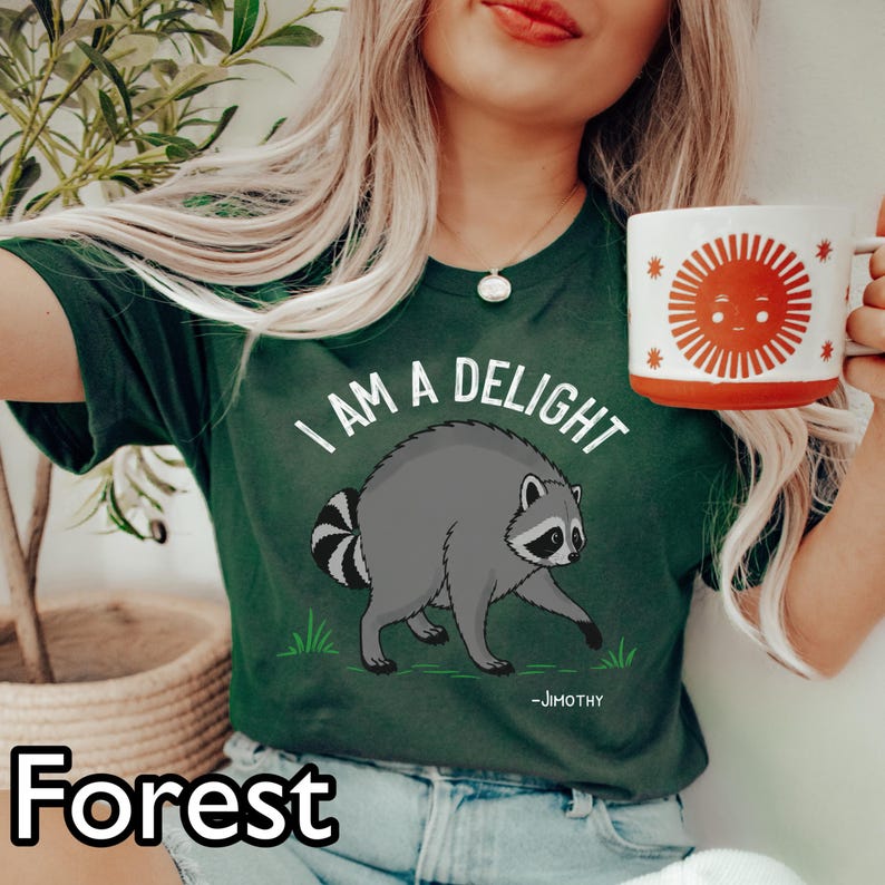 May include: A dark green t-shirt featuring a graphic of a gray raccoon with a striped tail, the text "I AM A DELIGHT" in white, and the name "-JIMOTHY" below. The shirt is paired with a white mug displaying a large orange sun illustration.