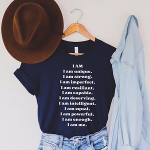 May include: A navy blue t-shirt with white text that reads "I AM I am unique. I am strong. I am imperfect. I am resilient. I am capable. I am deserving. I am intelligent. I am equal. I am powerful. I am enough. I am me."