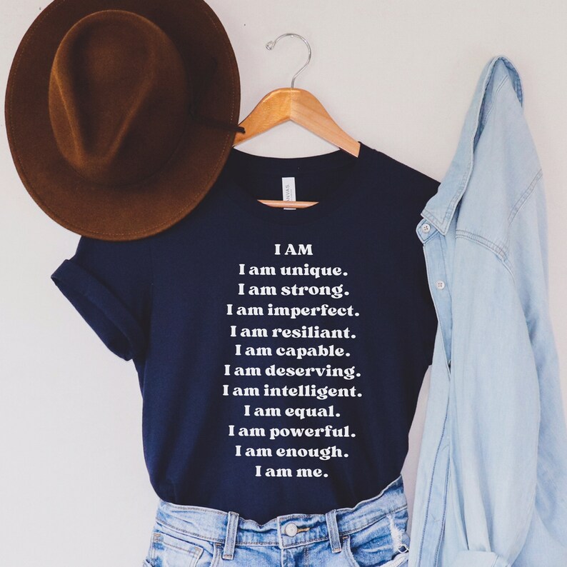 May include: A navy blue t-shirt with white text that reads "I AM I am unique. I am strong. I am imperfect. I am resilient. I am capable. I am deserving. I am intelligent. I am equal. I am powerful. I am enough. I am me."