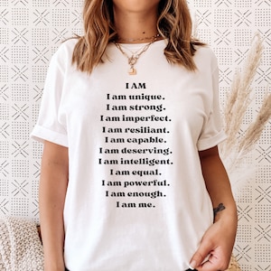 May include: White t-shirt with black text that reads "I AM I am unique. I am strong. I am imperfect. I am resilient. I am capable. I am deserving. I am intelligent. I am equal. I am powerful. I am enough. I am me."