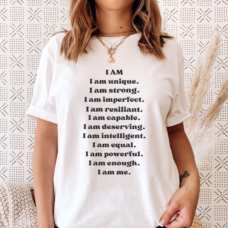 May include: White t-shirt with black text that reads "I AM I am unique. I am strong. I am imperfect. I am resilient. I am capable. I am deserving. I am intelligent. I am equal. I am powerful. I am enough. I am me."
