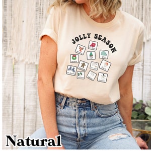 K&ouml;nnte beinhalten: Ein beiges T-Shirt mit dem Text "Jolly Season" in schwarzer Retro-Schrift. Das Shirt zeigt ein kreisf&ouml;rmiges Design aus wei&szlig;en Quadraten mit verschiedenen weihnachtlichen Illustrationen, darunter ein Rentier, ein Kranz, ein Keks, F&auml;ustlinge, Schlittschuhe, ein Schneemann und mehr.
