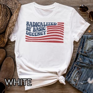 Pu&ograve; includere: T-shirt bianco con la scritta "RADICALIZED BY BASIC DECENCY" in blu e rosso, a forma di bandiera. La maglietta &egrave; annodata in basso. Sono visibili un cappello di paglia, occhiali da sole e jeans.