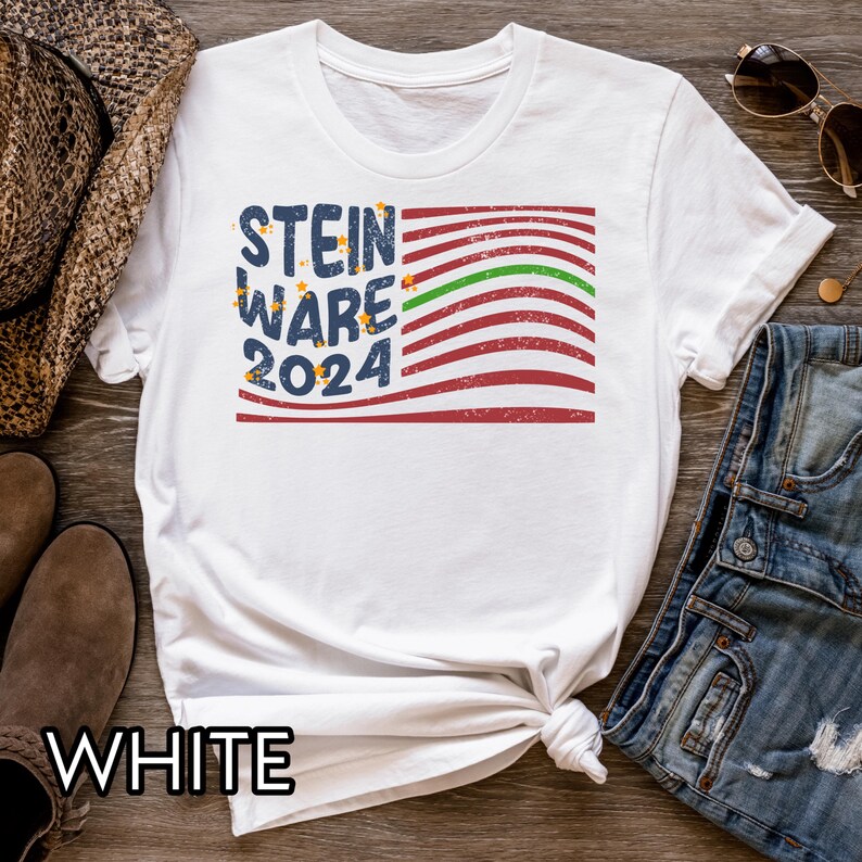 May include: White t-shirt with a red, white, and blue American flag design. The text "STEIN WARE 2024" is printed on the flag. The shirt is on a wooden surface with a pair of jeans and a cowboy hat.