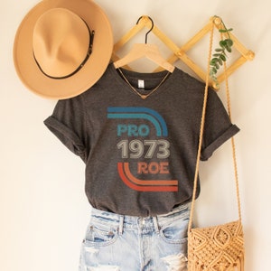 May include: A dark gray t-shirt with a vintage-style graphic that says "PRO 1973 ROE" in blue, red, and white. The graphic is surrounded by colorful stripes.
