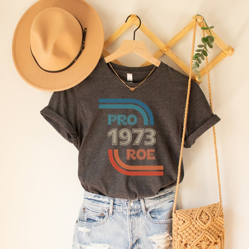 May include: A dark gray t-shirt with a vintage-style graphic that says "PRO 1973 ROE" in blue, red, and white. The graphic is surrounded by colorful stripes.