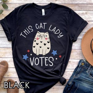 May include: Black t-shirt with a white graphic of a cat wearing glasses and the text "This Cat Lady Votes".