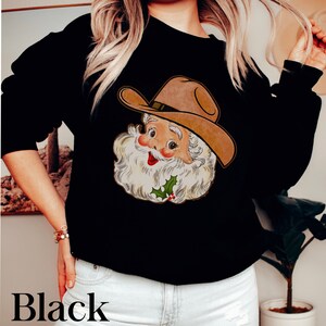 Puede incluir: Sudadera negra con una imagen de dibujos animados de Pap&aacute; Noel con un sombrero de vaquero. Pap&aacute; Noel sonr&iacute;e y tiene barba blanca y nariz roja.