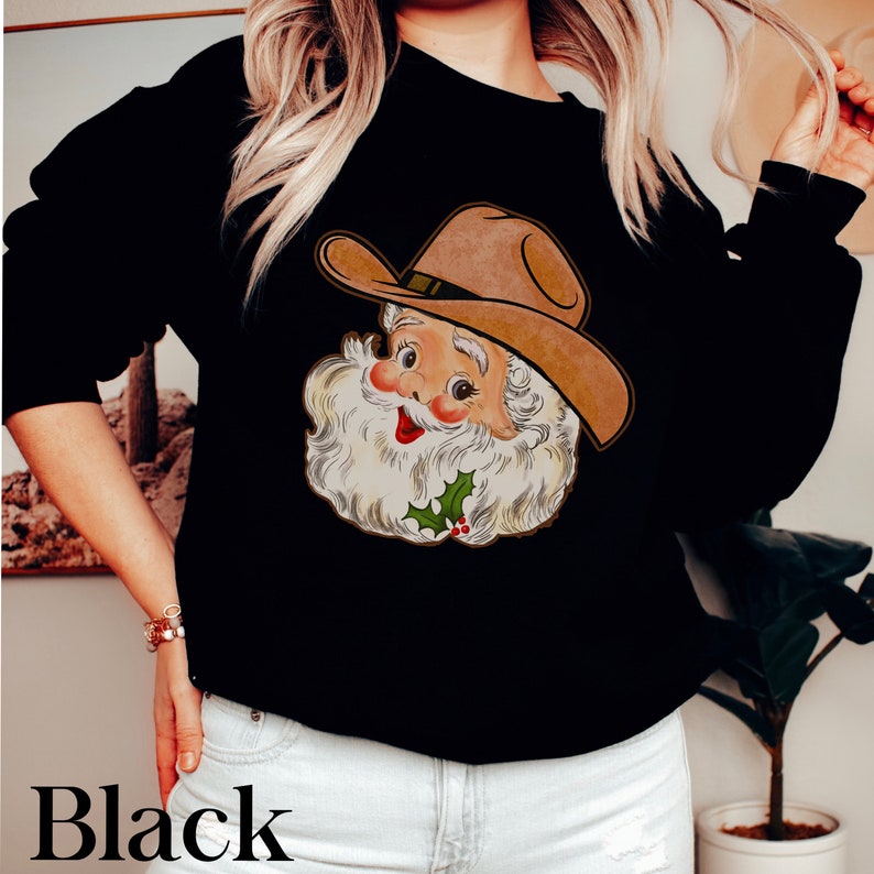 Puede incluir: Sudadera negra con una imagen de dibujos animados de Pap&aacute; Noel con un sombrero de vaquero. Pap&aacute; Noel sonr&iacute;e y tiene barba blanca y nariz roja.