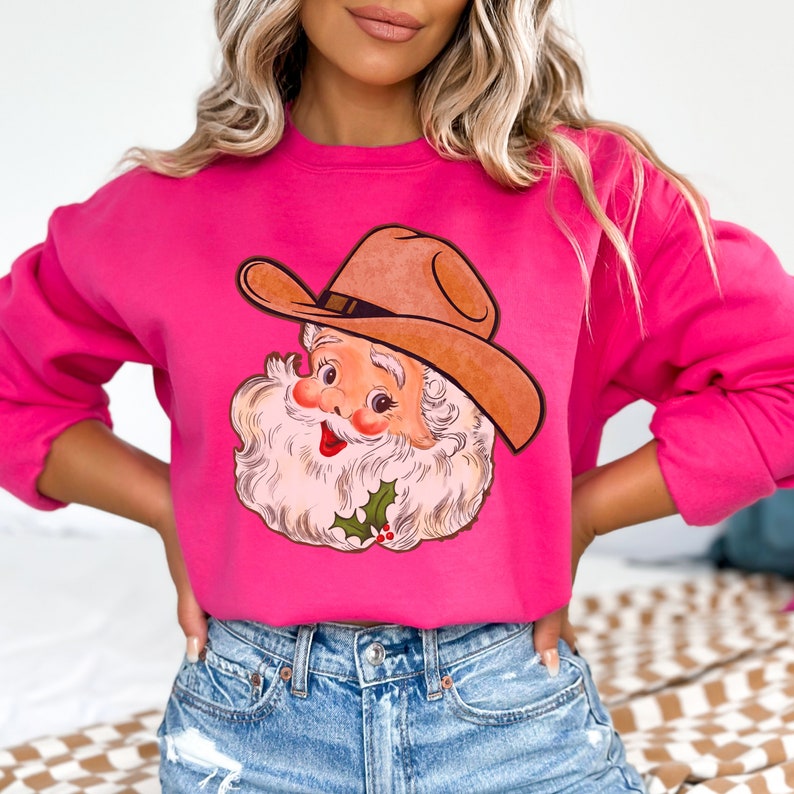 Puede incluir: Una sudadera rosa con una ilustraci&oacute;n vintage de Pap&aacute; Noel con un sombrero de vaquero. La ilustraci&oacute;n es de estilo de dibujos animados y presenta a un Pap&aacute; Noel sonriente con barba blanca y nariz roja.