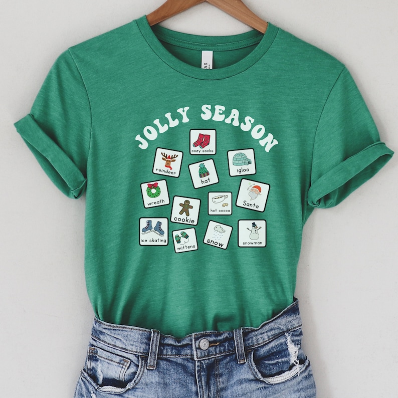 K&ouml;nnte beinhalten: Ein gr&uuml;nes T-Shirt mit dem Text "JOLLY SEASON" in einem wei&szlig;en Kreis. Im Kreis befinden sich 12 wei&szlig;e Quadrate mit Illustrationen zu Winterfeiertagsthemen, wie zum Beispiel ein Rentier, kuschelige Socken, ein Iglu, ein Hut, ein Kranz, der Weihnachtsmann, hei&szlig;e Schokolade, ein Keks, Schlittschuhlaufen, F&auml;ustlinge, Schnee und ein Schneemann.