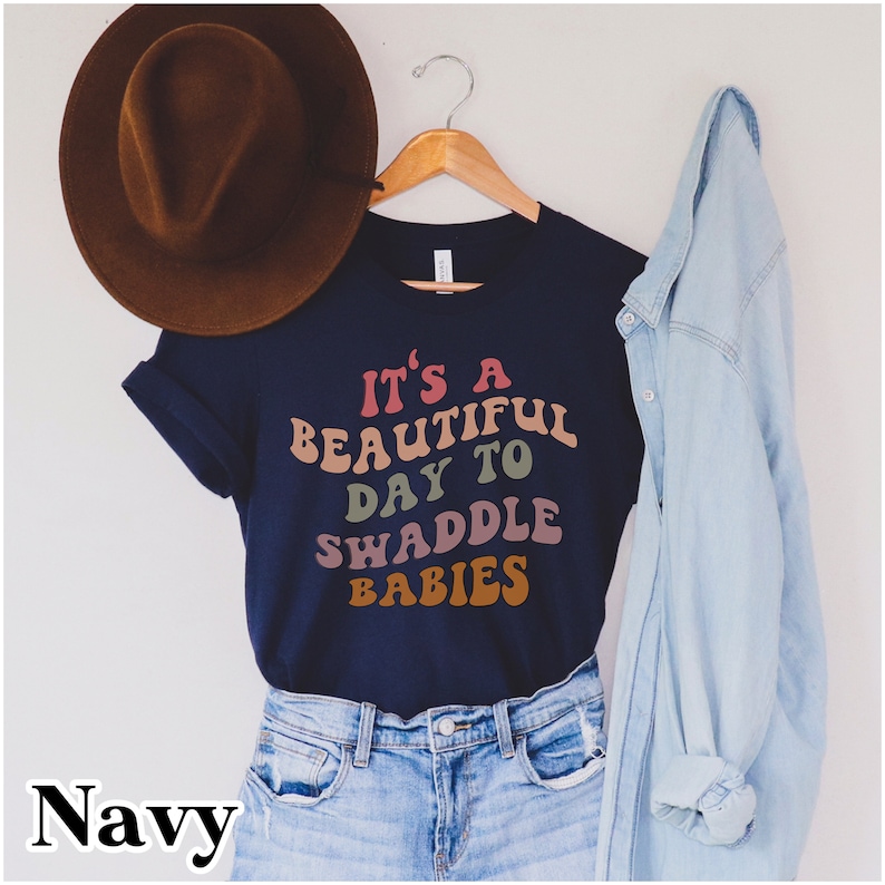 K&ouml;nnte beinhalten: Ein marineblaues T-Shirt mit dem Text "It's a beautiful day to swaddle babies" in Regenbogenfarben. Das Shirt h&auml;ngt an einem Kleiderb&uuml;gel und daneben liegen ein brauner Hut und eine blaue Jeansjacke.
