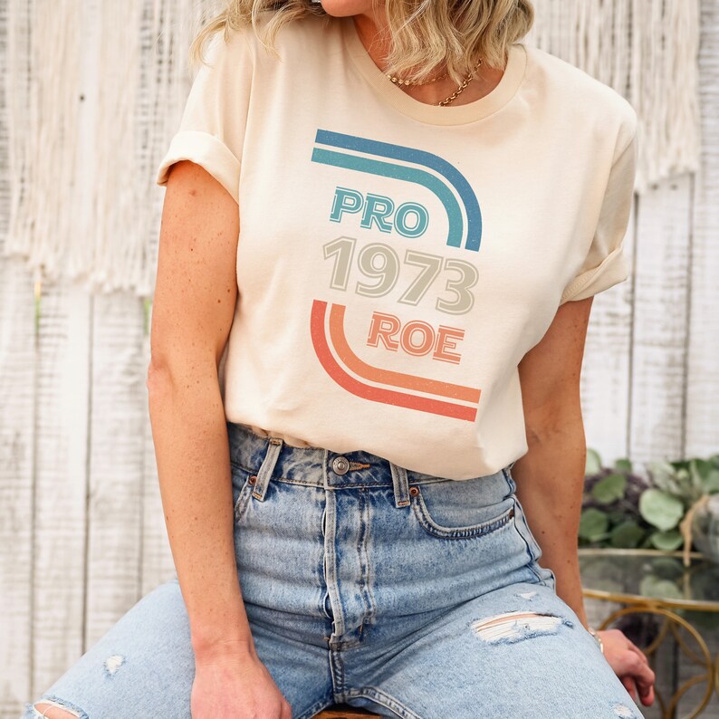 May include: A woman wearing a light beige t-shirt with a retro-style graphic that says "PRO 1973 ROE" in blue, white, and orange.