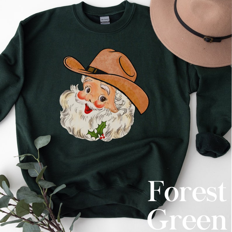 Puede incluir: Una sudadera de color verde bosque con una imagen de dibujos animados vintage de Pap&aacute; Noel con un sombrero de vaquero. El texto "Forest Green" est&aacute; impreso en la esquina inferior derecha de la sudadera.