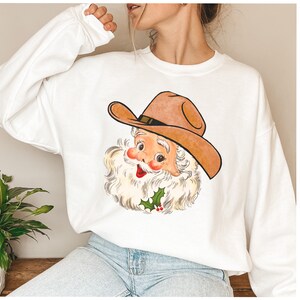 Puede incluir: Sudadera blanca con una imagen de dibujos animados de Pap&aacute; Noel con un sombrero de vaquero. Pap&aacute; Noel est&aacute; sonriendo y tiene una barba blanca. Hay una rama de acebo con bayas rojas debajo de su barbilla.