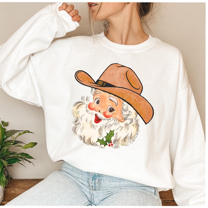 Puede incluir: Sudadera blanca con una imagen de dibujos animados de Pap&aacute; Noel con un sombrero de vaquero. Pap&aacute; Noel est&aacute; sonriendo y tiene una barba blanca. Hay una rama de acebo con bayas rojas debajo de su barbilla.