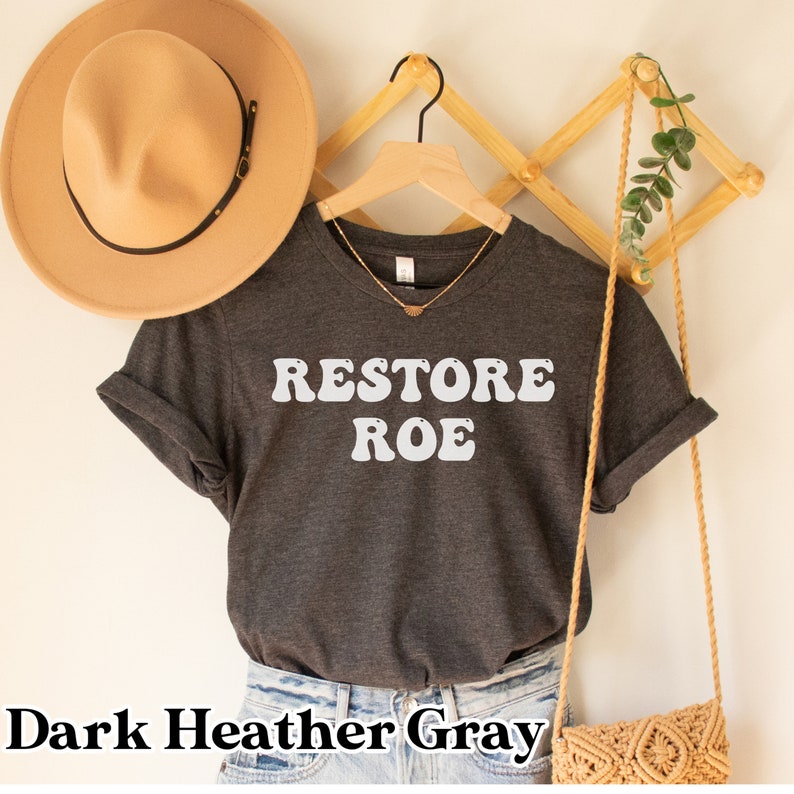 May include: Dark heather gray t-shirt with the text "RESTORE ROE" in white, retro-style lettering. The shirt is hanging on a wooden hanger.