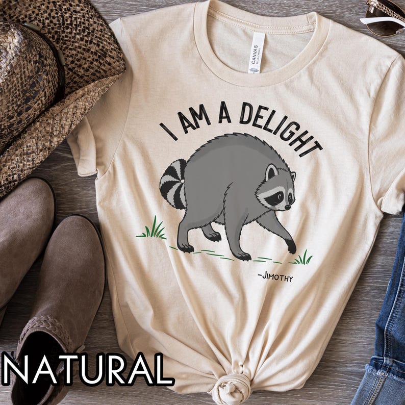 May include: A natural-colored t-shirt featuring a graphic of a gray raccoon with a striped tail. The text above the raccoon reads "I AM A DELIGHT" in black capital letters, and the name "-JIMOTHY" is printed in smaller black text below the raccoon.