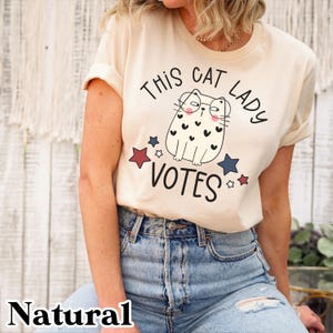 May include: A beige t-shirt with a graphic of a cat wearing glasses and the text "This Cat Lady Votes" with red and blue stars.