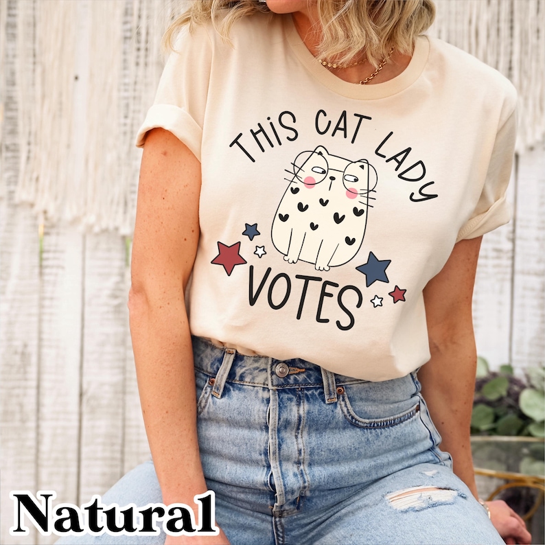 May include: A beige t-shirt with a graphic of a cat wearing glasses and the text "This Cat Lady Votes" with red and blue stars.