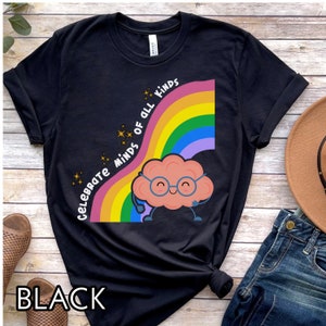Pu&ograve; includere: Maglietta nera con un arcobaleno e un cervello di cartone animato con gli occhiali. Il testo "Celebrate Minds of All Kinds" &egrave; sull'arcobaleno.