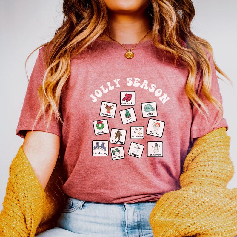 K&ouml;nnte beinhalten: Ein pinkfarbenes T-Shirt mit den Worten "Jolly Season" in einem wei&szlig;en Kreis. Im Kreis befinden sich 12 wei&szlig;e Quadrate mit Illustrationen von winterlichen Motiven, wie Rentier, kuschelige Socken, Iglu, Hut, Kranz, Keks, hei&szlig;e Schokolade, Weihnachtsmann, Schlittschuhlaufen, F&auml;ustlinge, Schnee und Schneemann.