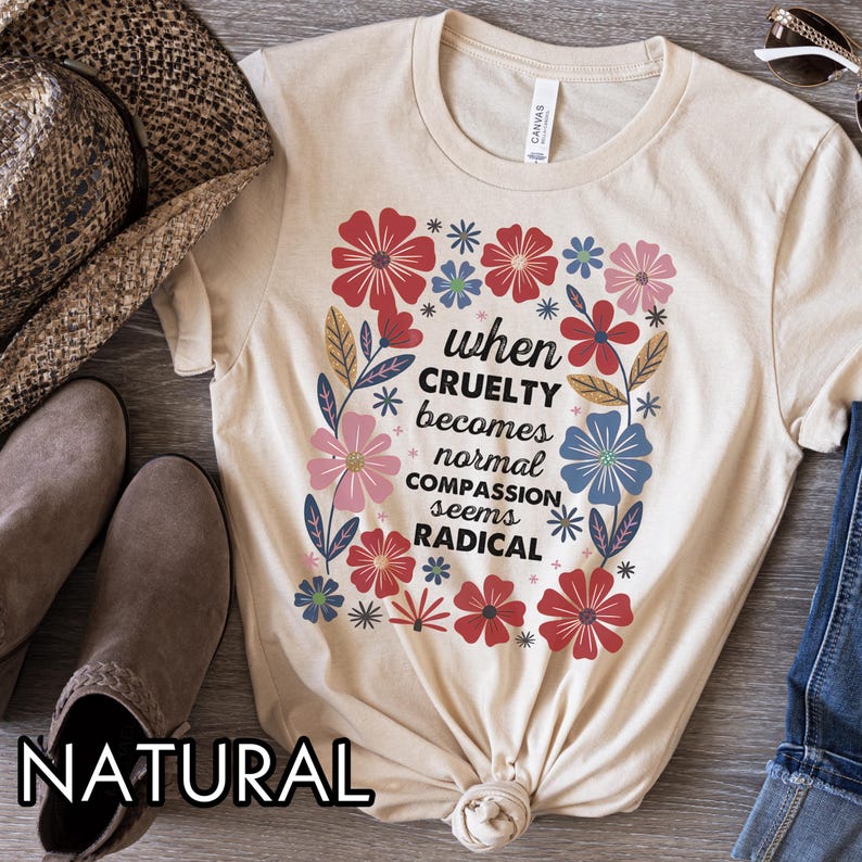 May include: A natural-colored t-shirt with the text "when cruelty becomes normal compassion seems radical" surrounded by colorful flowers. The flowers are red, pink, blue, and green. The shirt is knotted at the bottom.