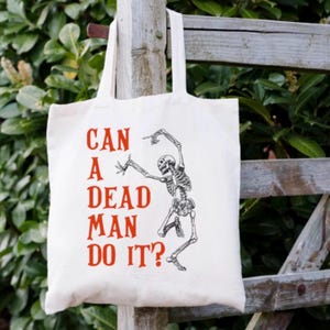 May include: A white canvas tote bag with red text that reads "Can a Dead Man Do It?" and a black and white illustration of a skeleton dancing.