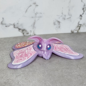 May include: A lavender-colored moth figurine with large, detailed wings. The wings feature a pink and white marbled design and are adorned with iridescent crystals. The moth has blue eyes and a fuzzy body, creating a whimsical aesthetic.