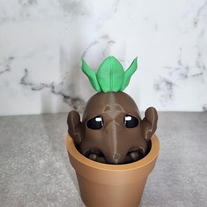 May include: A brown, 3D-printed mandrake figure with large, black eyes and small arms, emerging from a terracotta-colored flower pot. The mandrake has green leaves on top of its head. The pot sits on a gray surface against a marble-patterned background.
