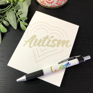 Pode incluir: Um cartão branco com um desenho de coração dourado e a palavra "Autismo" escrita em letras douradas. Uma caneta preta e prateada com a palavra "Autismo" escrita em cores do arco-íris está sobre o cartão.