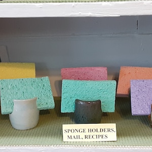 Puede incluir: Exhibición de coloridos porta esponjas con soportes de cerámica a juego. Las esponjas son de varios colores, incluyendo rosa, amarillo, verde, naranja, morado y azul. Los soportes de cerámica son en tonos verdes, blancos y marrones. El letrero dice "SPONGE HOLDERS, MAIL, RECIPES."