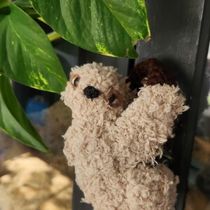 Puede incluir: Un juguete de peluche de perezoso hecho a mano en color beige claro. El perezoso tiene una nariz negra y ojos pequeños y oscuros. Sus brazos y pies son de un marrón más oscuro. El juguete está colocado cerca de una planta verde con hojas grandes.