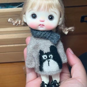 Op de afbeelding: Een kleine pop met blond haar in twee staartjes, gekleed in een grijze gebreide trui met een zwart-witte kattenafbeelding, een donkergrijze sjaal, witte legging en zwarte schoenen met oranje zolen, met grote donkere ogen en roze wangen.