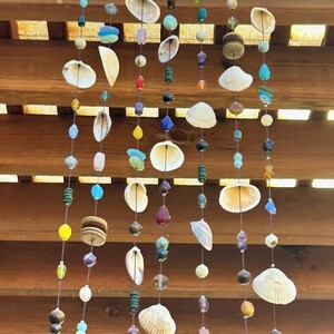 Puede incluir: Un móvil artesanal con conchas marinas, cuentas y piedras suspendidas de una rama de madera natural. Las cuentas son de color azul, verde, amarillo y marrón. Las conchas son blancas y beige. El móvil cuelga sobre un fondo de listones de madera.
