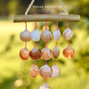May include: A seashell wind chime with a wooden bar and pink string. The chime features various seashells in shades of white, tan, and brown. The text "messy workshop ISLAND CREATIONS" is visible on the top.