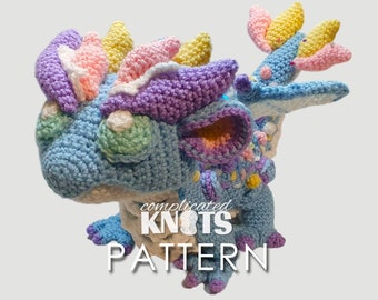 Könnte beinhalten: Ein farbenfrohes gehäkeltes Drachenspielzeug mit einem hellblauen Körper, lila und rosa Akzenten und einer gelben und rosa Mähne. Der Drache sitzt auf einem weißen Hintergrund mit dem Text "complicated KNOTS PATTERN" darunter.