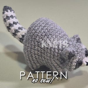 May include: A gray crocheted raccoon amigurumi toy with a white and gray striped tail. The toy has black eyes and a black nose. The toy is sitting on a light green surface. The text "PATTERN no sew!" is written below the toy.