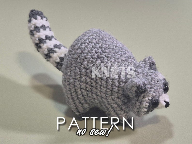 May include: A gray crocheted raccoon amigurumi toy with a white and gray striped tail. The toy has black eyes and a black nose. The toy is sitting on a light green surface. The text "PATTERN no sew!" is written below the toy.