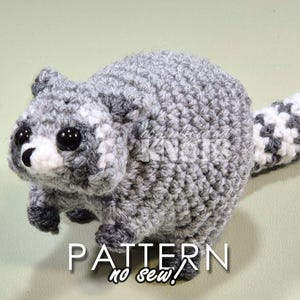 May include: A crocheted raccoon amigurumi toy with a gray and white striped tail. The toy has black eyes and a white nose. The toy is made with a no-sew pattern.