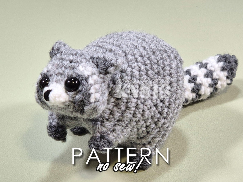 May include: A crocheted raccoon amigurumi toy with a gray and white striped tail. The toy has black eyes and a white nose. The toy is made with a no-sew pattern.
